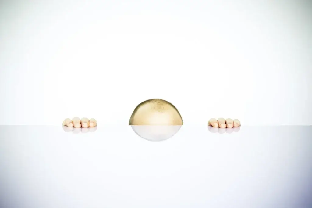 white table with white wall in background with bald man's fingers and top of head poking up from edge of table illustrating a buried penis