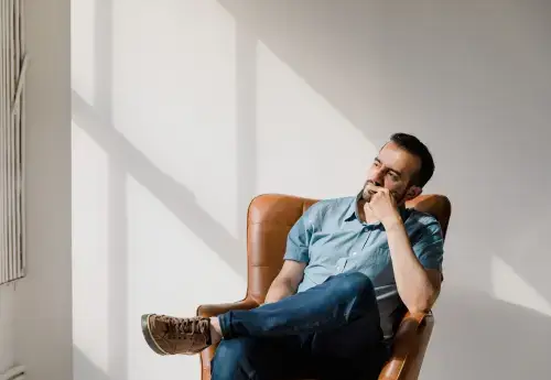a middle aged man wearing jeans and a blue shirt sits in a brown leather chair and peers out the window as he thinks about how he can get more girth