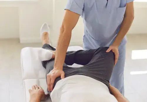 closeup of a medical professional positioning a man's legs as he lies on a table showing him how to perform male pelvic floor strengthening exercises