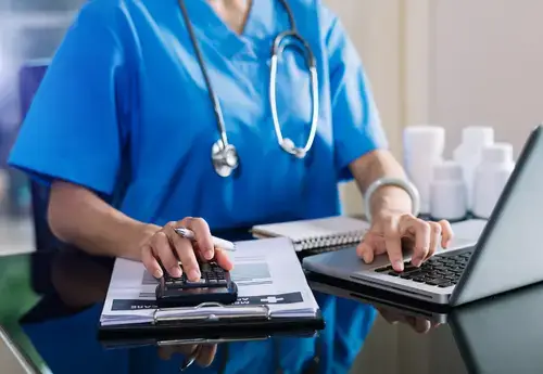 Healthcare professional calculating adult circumcision cost, using a laptop and calculator in a medical office. Financial planning and pricing considerations for adult circumcision procedures.