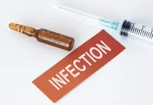 hypodermic needle in syringe and brown ampule filled with clear liquid next to a red sign that reads infection, signifying risk of infection from penuma implant