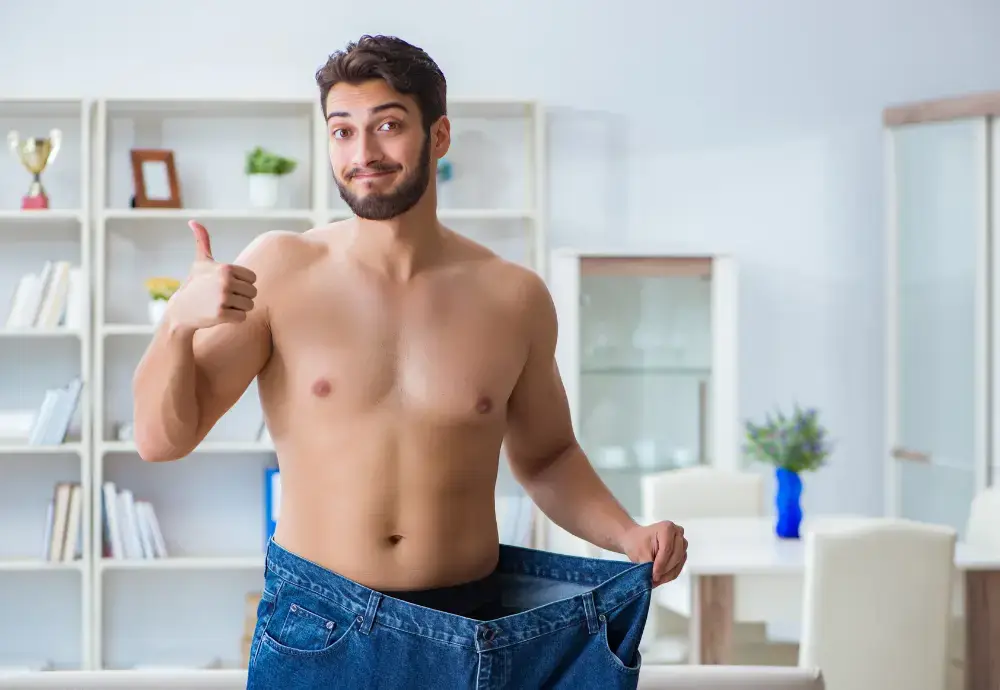 Shirtless man showing weight loss by wearing oversized jeans and giving a thumbs up — concept image for “does losing weight increase penis size.”