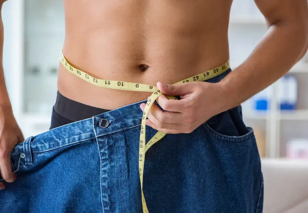 Man measuring waist with tape while wearing oversized jeans — visual representation for the question “does losing weight increase penis size.”