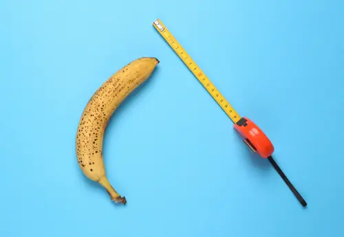 Ripe banana next to a tape measure on blue background, visually representing how to measure your penis length accurately.