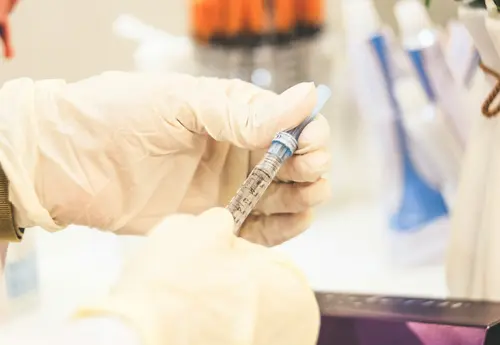 Close-up of medical professional preparing syringe for hyaluronic acid penile filler injection, with filler packaging visible in background.