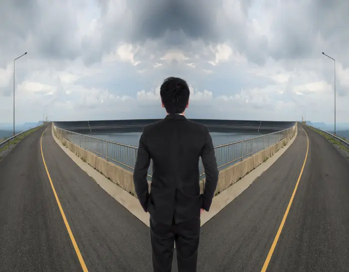 Man in a business suit standing at a forked highway looking at two diverging roads under a cloudy sky, symbolizing an important decision or choice.